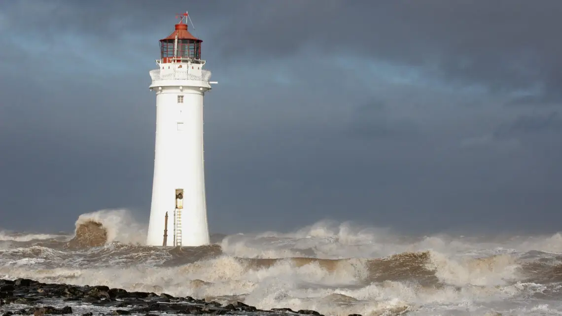 White lighthouse with a red top stands on a rocky shore as dark storm clouds loom and waves crash around it.